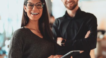 Lächelnde junge Frau mit Brille hält ein Tablet, im Hintergrund ein lächelnder junger Mann