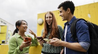 Eine Gruppe Studierende lacht im Freien mit Smartphones in den Händen.