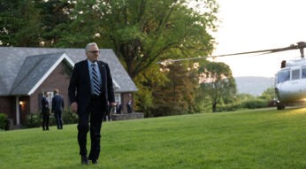 Auf dem Bild zur Serie Zero Day mit Robert De Niro ist Robert De Niro in einer eleganten schwarzen Anzug-Krawatten-Kombination zu sehen. Er geht über eine gepflegte Rasenfläche vor einem Backsteinhaus, während im Hintergrund ein weißer Helikopter steht. Die Szene wirkt angespannt und professionell, unterstützt durch die Anwesenheit von Sicherheitskräften im Hintergrund.