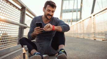 Mann sitzt auf dem Boden, macht Pause vom Sport und schaut lächelnd auf seine Smartwatch und sein Handy