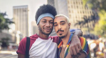 Auf dem Bild zur CSD Vodafone Kampagne umarmen sich zwei Männer und schauen selbstbewusst in die Kamera. Einer trägt ein Hemd in Regenbogenfarben, das Symbol für Pride und Vielfalt. Die Szene spielt in einer belebten Innenstadt mit Hochhäusern und sommerlicher Atmosphäre – ein Zeichen für Zusammenhalt, Sichtbarkeit und Offenheit.