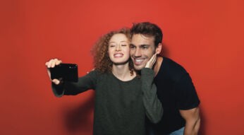 Zwei Personen machen ein Selfie von sich vor einem roten Hintergrund und freuen sich über das Freund:innen werben Programm von Vodafone