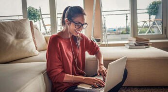 Auf dem Bild zum Internet für Studenten sitzt eine junge Frau mit Brille und dunklem Pferdeschwanz in einem roten Strickpullover entspannt auf einem Sofa und tippt lächelnd auf ihrem Laptop. Sie trägt weiße Kopfhörer und wirkt konzentriert, aber gut gelaunt. Im Hintergrund sorgen große Fenster mit Blick auf einen Balkon und ein Bücherstapel für eine helle, wohnliche und produktive Atmosphäre.
