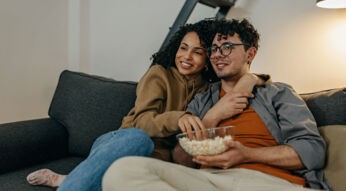 Mann und Frau sitzen lächelnd und kuschelnd auf dem Sofa mit einer Schüssel Popcorn in der Hand