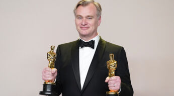 Christopher Nolan im Anzug hält in jeder Hand einen Oscar und guckt direkt in die Kamera.