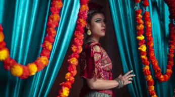 Ein junges Mädchen, traditionell gekleidet und geschminkt, blickt durch einen Vorhang, der mit Girlanden verziert ist.