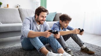 Ein Vater spielt mit seinem Sohn etwas auf einer Console und sitzt dabei auf dem Boden vor einer Couch