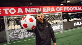 Podolski hält einen Fußball in die Kamera mit Vodafone Logo für die Baller League