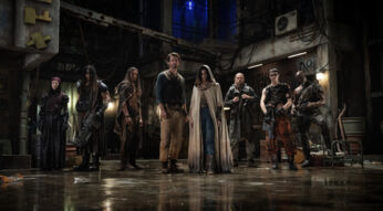 Der Rebel Moon Cast steht nebeneinander aufgereiht in einer alten Fabrikhalle