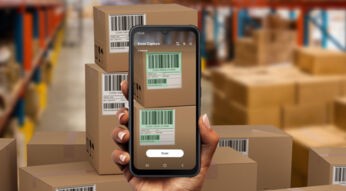 POV: Rechte Hand hält das robuiste Handy Galaxy Xcover7 in einer Lagerhalee vor einem Stapel Kartons