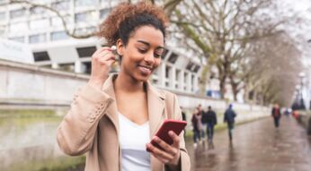 Eine junge Frau mit Smartphone in einer Hand. Mit der anderen Hand steckt sie einen In-Ear-Kopfhörer in ihr Ohr.