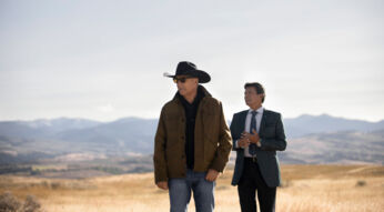 Auf dem Bild zum Faktenartikel über die Serie Yellowstone stehen Kevin Costner und Gil Birmingham in einer weiten Landschaft mit goldgelbem Gras und Bergen im Hintergrund. Kevin Costner trägt eine braune Jacke, Jeans, Sonnenbrille und einen schwarzen Cowboyhut, während Gil Birmingham einen eleganten grauen Anzug mit Krawatte trägt. Die Szene wirkt ruhig und nachdenklich, betont durch die offene Natur.