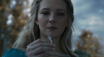 Auf dem Bild zu "Ringe der Macht Staffel 2" ist Galadriel, gespielt von Morfydd Clark, in einer Nahaufnahme zu sehen. Sie blickt konzentriert auf einen silbernen Gegenstand in ihrer Hand, während ihre blonden Haare im sanften Licht leuchten. Der Hintergrund ist leicht verschwommen, was den Fokus auf ihre ernste Miene und den geheimnisvollen Gegenstand lenkt.