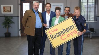 Der Cast von "Die Rosenheim-Cops" steht in einer Reihe und hält ein gelbes Ortseingangsschild mit "Rosenheim".