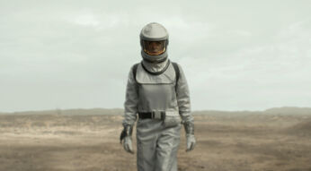 Auf diesem Bild aus Silo Staffel 2ist Rebecca Ferguson in einer beeindruckenden Wüstenlandschaft zu sehen. Sie trägt einen futuristischen Raumanzug in Silbergrau, der sie für die lebensfeindliche Umgebung außerhalb des Silos ausrüstet. Der Anzug ist mit einem dunkelgrauen Gurt und passenden Handschuhen ausgestattet. Ihr Helm verfügt über ein großes, klares Visier, das ihr Gesicht sichtbar macht und ihren ernsten, nachdenklichen Ausdruck hervorhebt. Der Hintergrund zeigt eine trostlose, weite Wüstenlandschaft, die eine karge und unwirtliche Atmosphäre vermittelt, passend zur dystopischen Thematik der Serie.