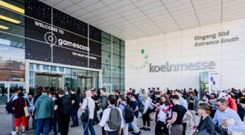 Das Bild zeigt den Eingang der Koelnmesse während der gamescom 2024. Vor dem Gebäude versammelt sich eine große Menschenmenge, die auf den Eintritt wartet. Über dem Eingang befindet sich ein großes Schild mit der Aufschrift "Welcome to gamescom - The Heart of Gaming". Die Stimmung ist lebhaft, und die Vorfreude auf das Event ist spürbar.