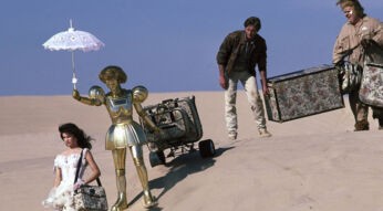 Ein Szenenbild aus Spaceballs zeigt Prinzessin Vespa, ihre Roboterin Dotty, Lone Starr und Waldi auf einer Wanderung durch die Wüste. Die Prinzessin geht voran, um ihre Schulter hängt eine TAsche. Hinter ihr geht die goldene Roboterin und hält einen Sonnenschirm über den Kopf der Prinzessin. Sie zieht einen Gepäckwagen hinter sich hier. Lone Starr folgt ihnen. Er hält mit einer Hand einen sperrigen Koffer. Auf der anderen Seite hilft Waldi beim Tragen des Koffers. Neben den Personen ist nicht als Sand und ein blauer Horizont zu sehen.