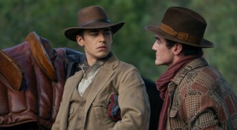 In dieser Szene aus Young Sherlock stehen sich Hero Fiennes Tiffin und Dónal Finn in einer intensiven Gesprächssituation gegenüber. Beide tragen breite Hüte und Outfits im Westernstil, im Hintergrund ist ein Sattel auf einem Pferd zu erkennen. Die Mimik der beiden wirkt ernst und angespannt, was auf eine dramatische Wendung hindeutet.