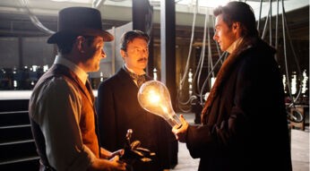 Drei Männer stehen in einer dunklen Werkstatt, einer von ihnen hält eine überdimensionierte Glühbirne in der Hand