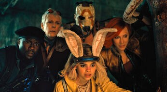 Im Bild zur Borderlands-Filmkritik sind 5 Hauptcharaktere zu sehen. Sie alle schauen erstaunt und neugierig in dieselbe RIchtung. Der Hintergrund ist dunkel, die Gesichter der Figuren werden von einer warmen Lichtquelle vor ihnen angestrahlt. Links im Bild befindet sich Roland (Kevin Hart). Erträgt eine dunkle Uniform mit schwarzer Soldaten-Kappe. Zentral im Bild und vor den anderen hockt Tiny Tina (Ariana Greenblatt). Sie hat eine gelbe Jacke an. Ihre Haare sind schulterlang und blond gefärbt. Auf ihrem Kopf trägt sie eine Art Haarreif mit plüschigen Hasenohren. Rechts im Bild ist Lilith (Cate Blanchett) zu sehen. Sie trägt dunkle Klamoten. Ihre Haare sind rot gefärbt. In der Mitte hinten steht Krieg (Florian Monteanue). Sein Körper ist von Tiny Tina verdeckt. Sein Gesicht ist ebenfalls nicht zu erkennen, denn er trägt eine weiße Maske mit einem orangenen Bogen darauf. Im Bild links von ihm ist Tannis (Jamie Lee Curtis) Gesicht zu sehen. Sie hat kurze graue Haare und trägt eine Art Sonnenbrille auf dem Kopf.