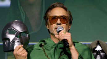 Auf dem Bild zu den Doctor Doom Fakten, welches auf der San Diego Comic Con 2024 entstand, hält Robert Downey Jr. eine silberne Doctor-Doom-Maske in der linken Hand und spricht in ein Mikrofon. Er trägt eine grüne Doctor-Doom-Kostüm mit Kette und hat eine orange getönte Sonnenbrille auf. Im Hintergrund ist ein weiterer Doctor-Doom-Darsteller mit Maske zu sehen.