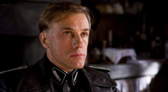 Auf dem Bild zu aus "Inglourious Basterds" ist eine der besten Szenen mit Christoph Waltz in seiner Rolle als SS-Standartenführer Hans Landa zu sehen. Er trägt eine schwarze SS-Uniform und blickt ernst und konzentriert nach vorne. Die Szene ist düster gehalten, mit einem unscharfen Hintergrund, der eine rustikale Innenumgebung andeutet.