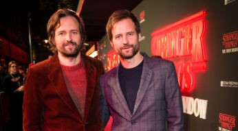 Auf dem Bild zu Something Very Bad Is Going To Happen stehen Matt und Ross Duffer, die Schöpfer von Stranger Things, nebeneinander vor einer Hintergrundwand mit dem Serienlogo zum Theaterstück "Stranger Things: The First Shadow". Matt Duffer trägt einen roten Cordanzug und einen passenden Pullover, während Ross Duffer einen grauen karierten Anzug mit einem dunklen T-Shirt kombiniert. Beide lächeln in die Kamera, während das rot beleuchtete Stranger Things Logo im Hintergrund leuchtet. Die Stimmung ist festlich und von Vorfreude geprägt.