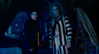 Auf diesem Bild zur Beetlejuice Beetlejuice Kritik stehen Winona Ryder als Lydia und Michael Keaton als Beetlejuice nebeneinander in einem düsteren, von Spinnweben bedeckten Dachboden. Lydia trägt schwarze Kleidung und blickt überrascht in die Kamera, während Beetlejuice in seinem markanten schwarz-weiß gestreiften Anzug grinst. Die Szene ist in kaltes, blaues Licht getaucht, was eine geheimnisvolle und leicht unheimliche Atmosphäre erzeugt. Im Hintergrund sind ein Leitergerüst und Abdeckungen zu erkennen.