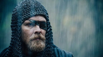 Auf dem Artikelbild von Hagen - Im Tal der Nibelungen ist die Figur Hagen in einer intensiven Szene bei Regen zu sehen. Er trägt ein Kettenhemd und eine schwarze Augenklappe, die sein ernstes, fokussiertes Gesicht betont. Sein wettergegerbtes, bärtiges Gesicht zeigt Entschlossenheit, während er in die Ferne blickt. Das Bild vermittelt eine düstere und kriegerische Atmosphäre, unterstrichen durch das graue, nasse Wetter.