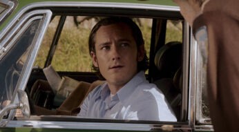 Auf dem Bild aus "Salems Lot - Brennen muss Salem" ist Lewis Pullman als Ben Mears zu sehen. Er sitzt in einem Auto, blickt nachdenklich und leicht besorgt nach oben zu einer Person, die außerhalb des Wagens steht. Seine Haltung und der ernste Ausdruck deuten auf eine angespannten Moment hin. Im Hintergrund sind grüne Bäume und Büsche zu erkennen, was die Szene ruhig wirken lässt.