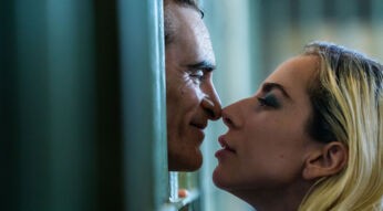 Auf diesem Bild von der *Folie à Deux*-Filmkritik sind Joaquin Phoenix als Joker und Lady Gaga als Harley Quinn in einer intensiven Szene zu sehen. Die beiden stehen sich sehr nah gegenüber, getrennt von Gefängnisgittern, mit nur wenigen Zentimetern Abstand zwischen ihren Gesichtern. Phoenix lächelt leicht, während Lady Gaga ihn mit halbgeschlossenen Augen ansieht, bereit, ihn fast zu küssen. Die Szene strahlt Spannung und Nähe aus, unterstützt durch das gedämpfte Licht und die dunklen Farben im Hintergrund.
