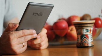 Hände halten einen Kindle-eReader. Auf dem Tisch steht eine bunte Tasse, im Hintergrund eine Obstschale mit Äpfeln gefüllt.