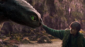 Auf dem Bild zur neuen Verfilmung von Drachenzähmen leicht gemacht für 2025 ist der schwarze Drache Ohnezahn mit grünen Augen links zu sehen, wie er sanft mit Hicks interagiert. Hicks, rechts im Bild, trägt grüne Kleidung mit einer Fellweste und berührt die Nase von Ohnezahn. Die Szene spielt in einer naturbelassenen, waldigen Umgebung mit gedämpften Farben, die eine ruhige und emotionale Atmosphäre schaffen.