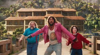 Auf dem Bild aus Ein Minecraft Film sind Jack Black als Steve, Jason Momoa als Garrett und Sebastian Hansen als Henry zu sehen. Sie wirken überrascht und alarmiert, während sie auf einer Brücke vor einer detailreichen, blockartigen Minecraft-Landschaft stehen. Im Hintergrund erhebt sich ein großes Haus im typischen Minecraft-Stil mit grünen Bäumen und Bergen.