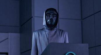 Das Bild im Artikel zum Squid Game US-Remake zeigt eine Szene aus der zweiten Staffel der Originalserie, in der der Frontman, eine mysteriöse Figur in einem kantigen, schwarzen, geometrischen Maske und einem grauen Kapuzenmantel, hinter einem Pult steht, umgeben von einer düsteren, blau beleuchteten Umgebung.