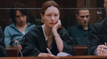 Auf dem Bild in dem Artikel über die Zeit Verbrechen-Serie, ist eine Frau mit roten Haaren zu sehen, die wie eine Anwältin gekleidet ist. Im Hintergrund sind weitere Menschen zu sehen. Der Raum in dem sie sitzen scheint ein Gerichtssaal zu sein.