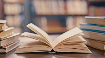 Ein Buch liegt aufgeklappt auf einem Tisch zwischen zwei Bücherstapeln.