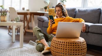 Frau mit Kopfhörern sitzt auf dem Boden vor einer Couch und hört Musik via Smartphone.