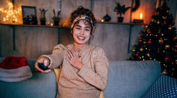 Eine lachende Frau sitzt gemütlich auf einem Sofa und schaut Weihnachtsepisoden von Serien