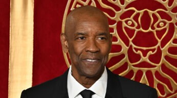 Auf dem Bild zu Denzel Washingtons besten Filmmomenten ist Denzel Washington in einem eleganten schwarzen Anzug mit weißem Hemd und schwarzer Krawatte zu sehen. Er lächelt dezent, während er vor einem roten Hintergrund mit goldenen Verzierungen, die ein Löwenmotiv zeigen, steht. Die Szene strahlt eine feierliche und stilvolle Atmosphäre aus.