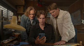 Bob Andrews (Levi Brandl), Peter Shaw (Nevio Wendt) und Justus Jonas (Julius Weckauf) in einer Szene aus "Die drei ??? und der Karpatenhund". Die drei Detektive stehen in ihrem Büro, vertieft in das Display eines Smartphones, das Peter hält.