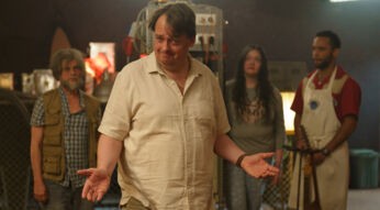 Auf dem Bild aus der Serie auf Amazon Prime Video ist Sascha Nathan in der Rolle des Gerry Star zu sehen. Gerry steht im Mittelpunkt einer Gruppe von vier Personen, die ihn aus dem Hintergrund beobachten. Er trägt ein beiges Hemd und blickt mit einem schelmischen Lächeln in die Kamera, seine Arme lässig ausgebreitet. Im Hintergrund sind Laborgeräte und eine industrielle Umgebung zu erkennen. Die Szene wirkt humorvoll und leicht chaotisch.