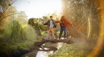 Auf dem Visual zum Weihnachtsspot von Vodafone 2024 balancieren zwei Jugendliche fröhlich auf einem umgestürzten Baumstamm über einen kleinen Bach in einer idyllischen Naturkulisse. Sie tragen lässige, bunte Kleidung, während goldenes Sonnenlicht durch die Bäume scheint und eine warme, festliche Stimmung erzeugt.