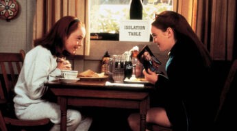 Auf dem Bild aus Lindsay Lohans Film Ein Zwilling kommt selten allein, einem ihrer besten Filme, sind die Zwillinge Hallie und Annie zu sehen, beide gespielt von Lindsay Lohan. Sie sitzen an einem Holztisch mit der Aufschrift "Isolation Table" und unterhalten sich lebhaft. Hallie trägt einen grauen Pullover, während Annie eine dunkle Jacke trägt und ein Foto in der Hand hält. Im Hintergrund sieht man ein Fenster, durch das Tageslicht einfällt.