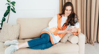 Eine junge Frau sitzt auf einem Sofa und blickt auf ihr Smartphone.