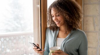 Frau schaut lächelnd auf ihr Android-Smartphone, während sie eine Tasse Tee in der Hand hält.