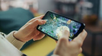Auf dem Bild ist eine Person zu sehen, die ein Smartphone in beiden Händen hält und ein Strategiespiel spielt. Der Bildschirm zeigt eine farbenfrohe Spielszene mit Figuren und Steinelementen. Die entspannte Umgebung im Hintergrund deutet auf Freizeit hin. Das Bild thematisiert Handyspiele für Erwachsene.