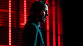 Auf dem Bild in dem Artikel zu den John Wick-Zitaten ist John Wick, gespielt von Keanu Reeves, in einem dunklen Anzug zu sehen. Er steht im Profil vor einem Hintergrund mit leuchtend roten Lichtern. Sein Blick ist ernst und fokussiert, die Szene wirkt stilisiert und atmosphärisch düster, typisch für die Ästhetik der John Wick-Filme.