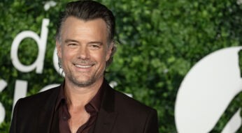 Auf dem Bild ist Josh Duhamel zu sehen, der lächelnd vor einer Kulisse mit grünem Laub und verschwommenem Schriftzug steht. Er trägt einen eleganten dunkelbraunen Anzug mit passendem Hemd und wirkt entspannt und charmant. Das Bild ist Teil einer Präsentation oder Veranstaltung und unterstreicht den Stil des Schauspielers.
