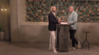 In diesem Bild aus „Promi First Dates“ ist Paul Janke zu sehen, der in einem dunklen Sakko, einem weißen Hemd und weißen Hosen an einem Tresen steht. Er unterhält sich gestikulierend mit einem Mann in einem hellblauen Sakko und weißen Sneakern. Im Hintergrund ist eine Wand mit einer bunten, geometrischen Dekoration zu erkennen, die dem Raum eine moderne Atmosphäre verleiht.